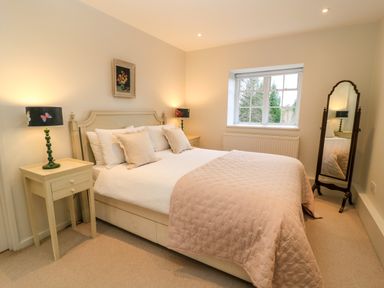 A bedroom with a large bed, two bedside tables with lamps, a window, a mirror, and a painting above the bed at The Elm Coach House in Chipping Norton