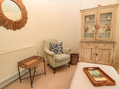 A corner of a room with an armchair, a wicker table, a wooden cabinet with glass doors, a mirror on the wall, and a wooden tray with a book on a bed at The Elm Coach House in Chipping Norton