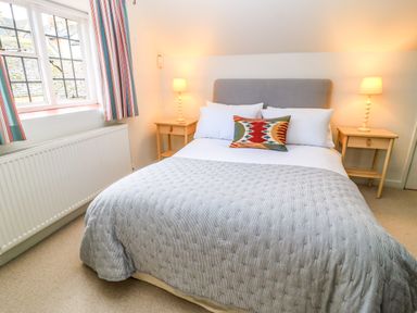 A bedroom with a double bed grey headboard patterned pillow two wooden bedside tables with lamps window with striped curtains and radiator at The Elm Coach House in Chipping Norton