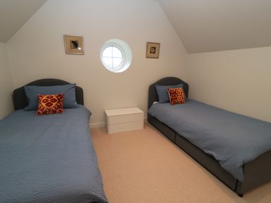 A bedroom with two single beds with blue bedding and patterned cushions a small white chest of drawers and a circular window at The Elm Coach House in Chipping Norton