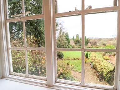 A view of a garden with grass bushes and trees through a white framed multi pane window at The Elm Coach House in Chipping Norton