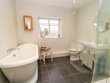 A bathroom with a freestanding bathtub a chair with towels a toilet a sink cabinet a round mirror and a window at The Elm Coach House in Chipping Norton