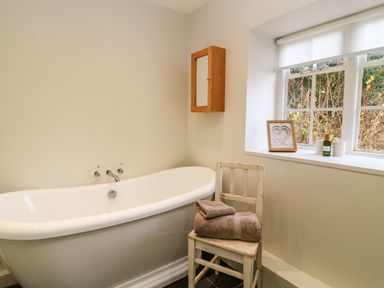 A bathroom with a freestanding bathtub a wooden chair with folded towels a wooden cabinet and a window with a framed picture and toiletries at The Elm Coach House in Chipping Norton