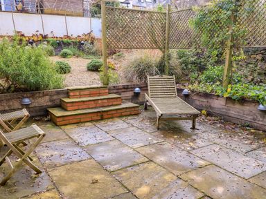 An outdoor patio with stone flooring a wooden lounge chair wooden chairs brick steps and garden beds at The Elm Coach House in Chipping Norton