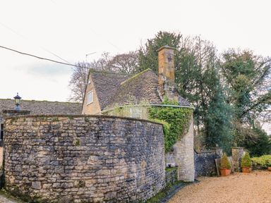 A stone house with a curved stone wall and trees in the background at The Elm Coach House in Chipping Norton