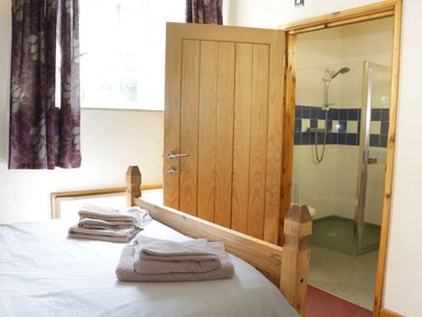 A bedroom with a wooden bed and folded towels on the bed next to a door leading to a tiled bathroom with a shower at Woodside Cottage in Kerry