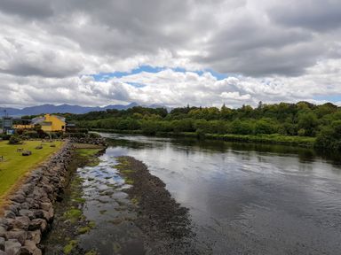 A river flowing beside a grassy area with a yellow building and mountains in the background at Knockbrack Lonart near Killorglin County Kerry
