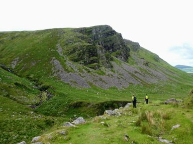 Two hikers walking on a grassy trail near a rocky hillside in a mountainous area at Knockbrack Lonart near Killorglin County Kerry