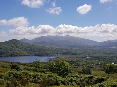 A landscape with a lake surrounded by green hills and trees with mountains in the background at Knockbrack Lonart near Killorglin County Kerry