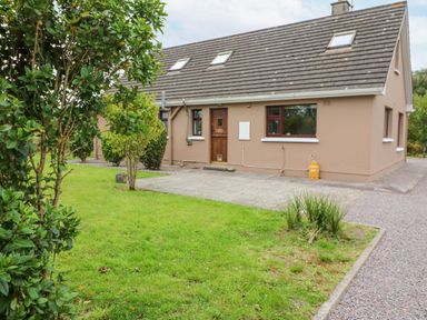 A brown house with a tiled roof a wooden door and multiple windows surrounded by grass and bushes at Knockbrack Lonart near Killorglin County Kerry