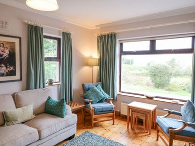 A living room with beige sofa blue armchairs small wooden tables and large windows at Knockbrack Lonart near Killorglin County Kerry