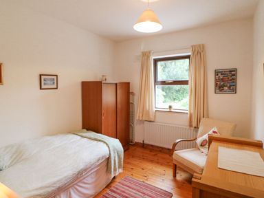 A bedroom with a single bed, wooden wardrobe, armchair, desk, and window with curtains at Knockbrack in Lonart near Killorglin County Kerry