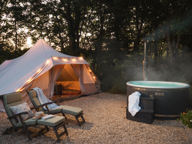 A tent with string lights beside two padded lounge chairs and a hot tub in a wooded outdoor area at Moonshine in St Erme near Truro