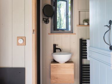 A bathroom with a round white sink on a wooden cabinet a black faucet a small window a wall-mounted round mirror a towel on a black radiator and a plant on a wall shelf at Moonshine in St Erme near Truro