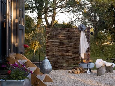 An outdoor area with wooden steps leading to a door fire pit with stacked wood and a hat and cloth on a wooden fence at Moonshine in St Erme near Truro