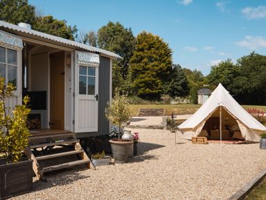 An open doorway of a small gray building with steps and potted plants next to a white canvas tent on a gravel area with trees and grass in the background at Stargazer in St Erme near Truro