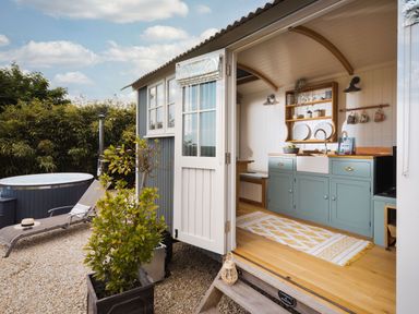 An open door showing a compact kitchen area with green cabinets and wooden shelves next to an outdoor area with a lounge chair and a hot tub at Stargazer in St Erme near Truro