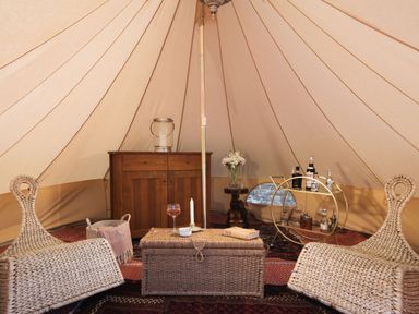 A tent interior with two wicker chairs a wicker chest a wooden cabinet a small table with flowers and a round bar cart with bottles at Stargazer in St Erme near Truro