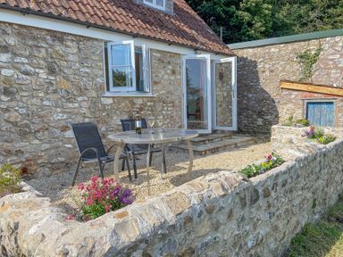 An outdoor patio area with a round table, two chairs, flowers in stone planters, and a stone building with open doors and windows at The Wheelhouse Beacon near Yarcombe