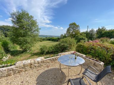 An outdoor patio with a metal table holding wine glasses and a bottle surrounded by chairs and overlooking a garden at The Wheelhouse Beacon near Yarcombe