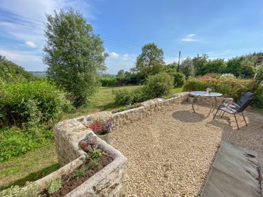 A gravel patio with a metal table and two chairs overlooking a garden and trees at The Wheelhouse Beacon near Yarcombe