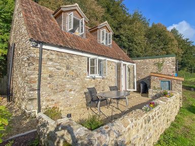 Exterior of a stone cottage with two dormer windows a small patio with table and chairs and a stone wall at The Wheelhouse Beacon near Yarcombe