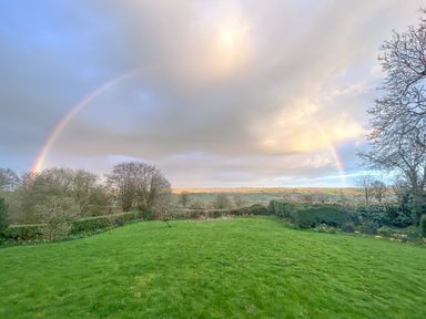 A green lawn with trees and bushes under a cloudy sky with a rainbow at The Wheelhouse in Beacon near Yarcombe