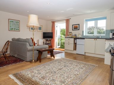 An open living space with a gray sofa a wooden bench a floor lamp a television a kitchen with cream cabinets and a window at The Wheelhouse Beacon near Yarcombe