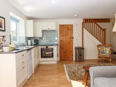 A kitchen with white cabinets and appliances next to a wooden door and staircase with a wicker chair and rug at The Wheelhouse in Beacon near Yarcombe