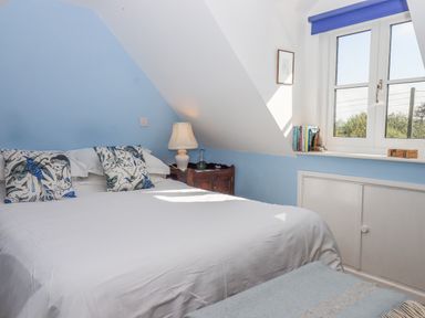 A bedroom with a bed white bedding and patterned cushions a lamp on a wooden nightstand books on a windowsill and blue walls at The Wheelhouse Beacon near Yarcombe