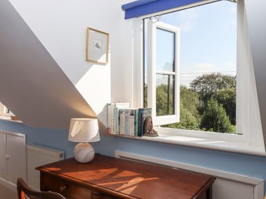 A wooden desk with a lamp and books on the windowsill under an open window in a room with slanted ceiling at The Wheelhouse Beacon near Yarcombe