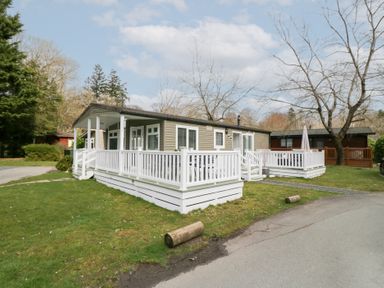 A single story cabin with white railings on a porch and surrounding grass at Cosy Corner in White Cross Bay near Troutbeck Bridge