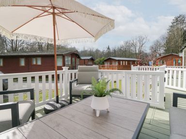 An outdoor patio with a table and chairs under a large umbrella surrounded by white fencing and wooden cabins in the background at Cosy Corner White Cross Bay near Troutbeck Bridge