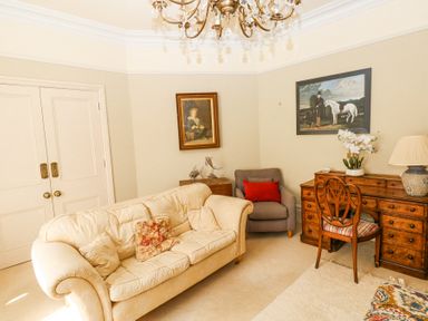 A sitting room with a cream sofa and floral cushions a gray armchair with a red cushion a wooden desk with a chair and lamp and two paintings on the walls at Hereford House in Tenby