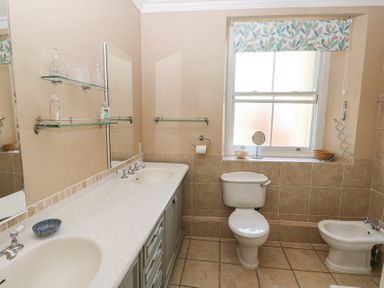 A bathroom with a double sink countertop toilet bidet window with floral valance shelves with bottles and small mirror