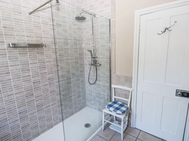 A bathroom with a glass shower enclosure tiled walls and a white chair with a blue checkered cushion near a white door