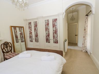 A bedroom with a bed and two rolled towels a wooden chair a full-length mirror built-in wardrobes with floral curtains an arched doorway and a window with curtains at Hereford House in Tenby
