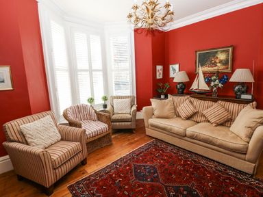 A living room with red walls, a beige sofa, striped chairs, a wicker chair, a patterned rug, lamps, and framed artwork at Hereford House in Tenby