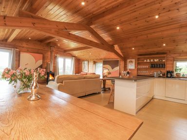 An open plan living area with a wooden ceiling and beams a beige sofa a wood burning stove and a kitchen with cream cabinets at Artisan Lodge in Longridge