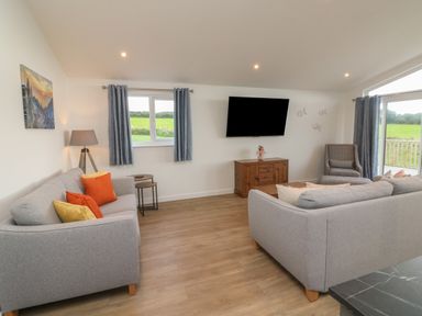 A living room with two gray sofas, a wooden cabinet with a TV mounted above it, a patterned armchair, and windows with blue curtains at Bay in Ilfracombe