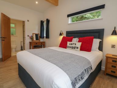 A bedroom with a large bed with red pillows and a white pillow with text a wooden dressing table and an open door leading to a bathroom at Bay in Ilfracombe