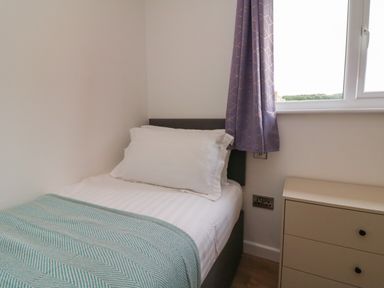 A single bed with white pillow and blanket next to a window with purple curtains and a beige chest of drawers at Bay in Ilfracombe