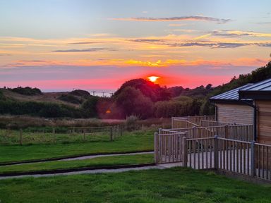 A sunset over grassy fields with bushes and a building with a wooden deck in the foreground at Cove Lodge in Ilfracombe