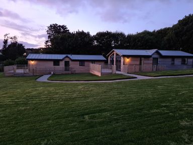Two wooden lodges with porches surrounded by grass and trees at dusk at Cove Lodge in Ilfracombe