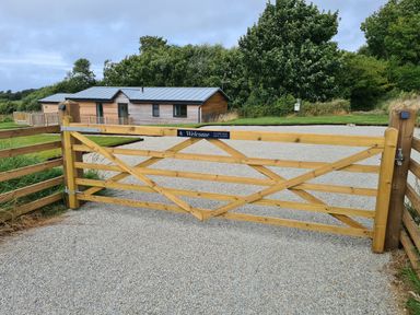 A wooden gate across a gravel driveway leading to a wooden house with a metal roof surrounded by trees at Cove Lodge in Ilfracombe
