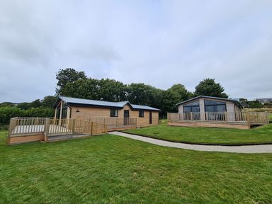 Two wooden lodges with decks on a grassy lawn with trees in the background at Cove Lodge in Ilfracombe