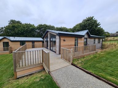 A wooden lodge with large glass doors and a fenced wooden deck surrounded by grass at Cove Lodge in Ilfracombe