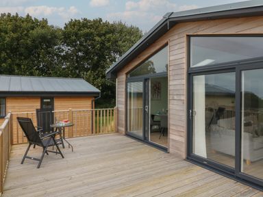 A wooden deck with two black chairs and a small round table outside a wooden house with large glass doors at Cove Lodge in Ilfracombe