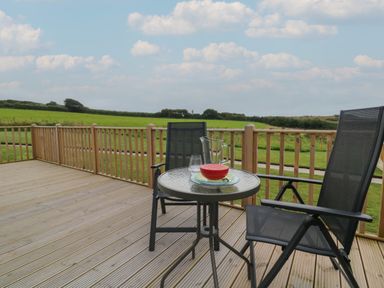 A wooden deck with a round glass table and two black chairs overlooking green fields at Cove Lodge in Ilfracombe