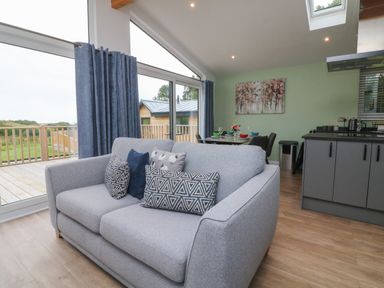 A sitting area with a gray sofa and cushions next to large windows and a dining table with chairs at Cove Lodge in Ilfracombe